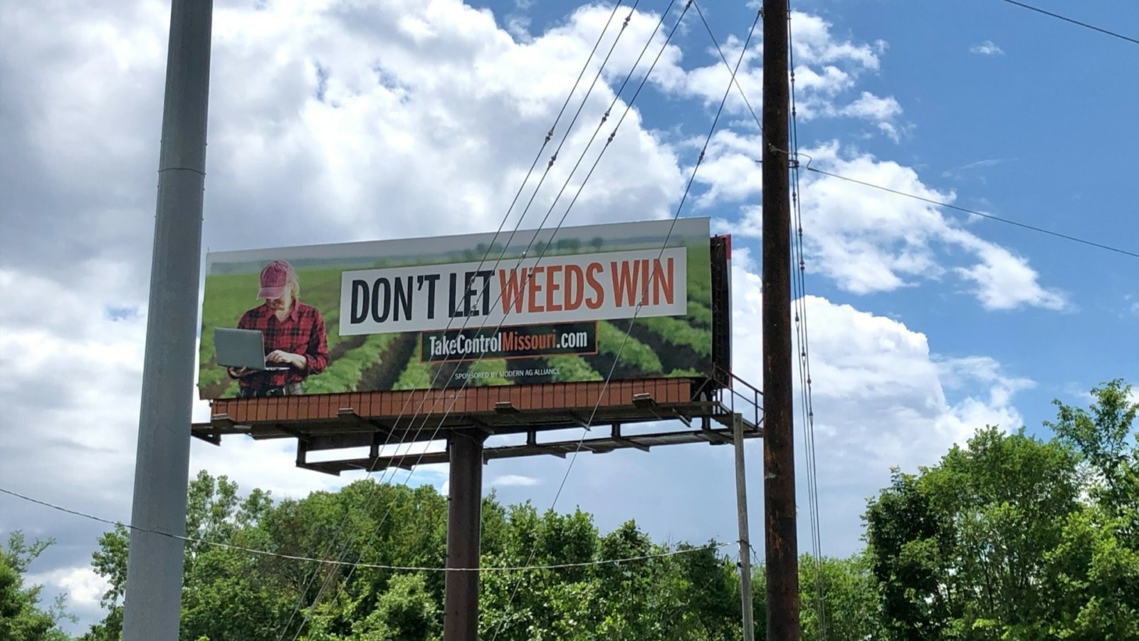 A billboard with a farmer holding a laptop in rows of a crop. Text on the billboard reads "Don't let weeds win" and "takecontrolmissouri.com"