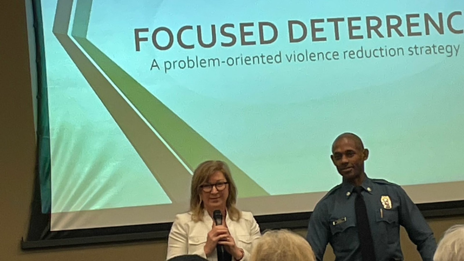 A man in a police uniform and a woman in a white blazer stand before a screen with the words "Focused Deterrence."