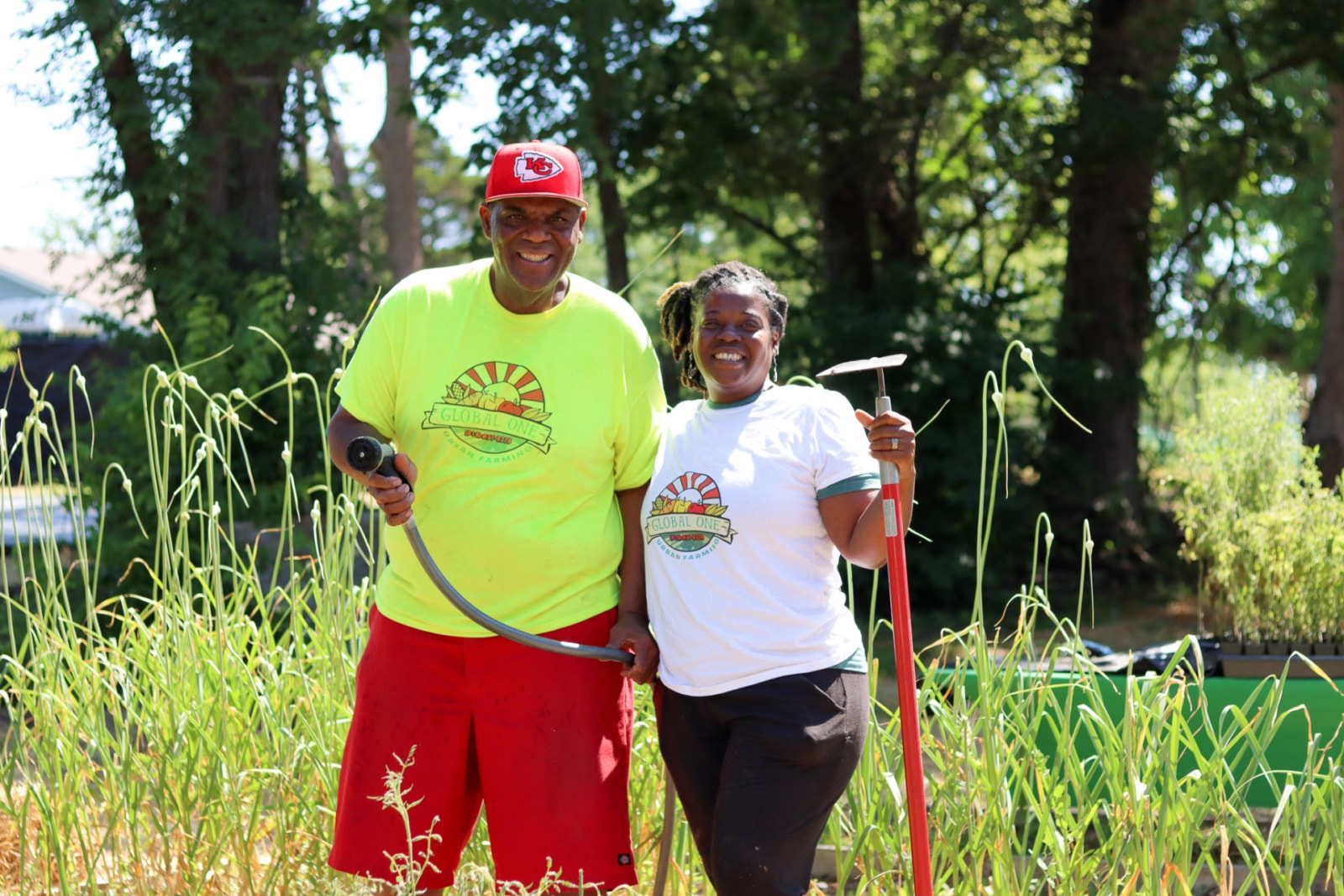 a man in a red hat, yellow shirt and red shorts holds up a garden hose and stands in front of a bed of garlic plants, next to a woman in a white shirt and black pants who holds up a garden hoe. Both of their shirts read "Global One urban farming"