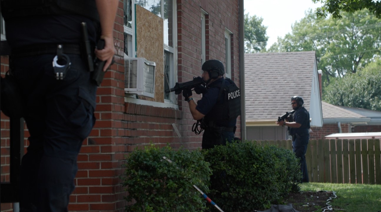 A Police Officer in a bulletproof vest with a high-powered assault rifle stares into the ground-level window of a residence. There are two other officers on either side of him.