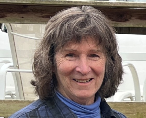 A women with a blue turtleneck and dark jacket with dark hair smiles in front of an indiscernible background.