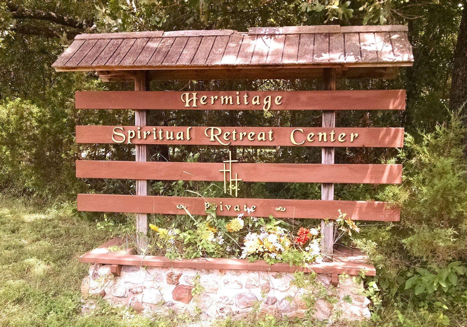 A large wooden sign in a treed outdoor setting. The sign says "Spiritual Hermitage Retreat Center" in gold scripted lettering.