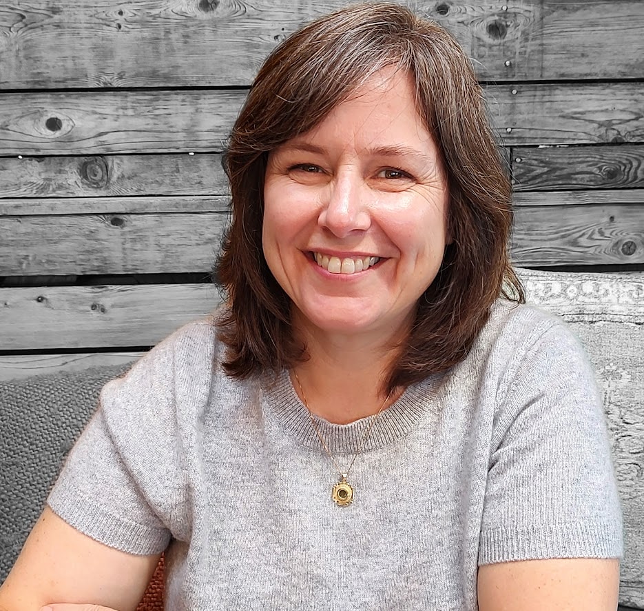 A woman with brown hair and a gray t-shirt sits smiling in front of a wall of weathered wood.